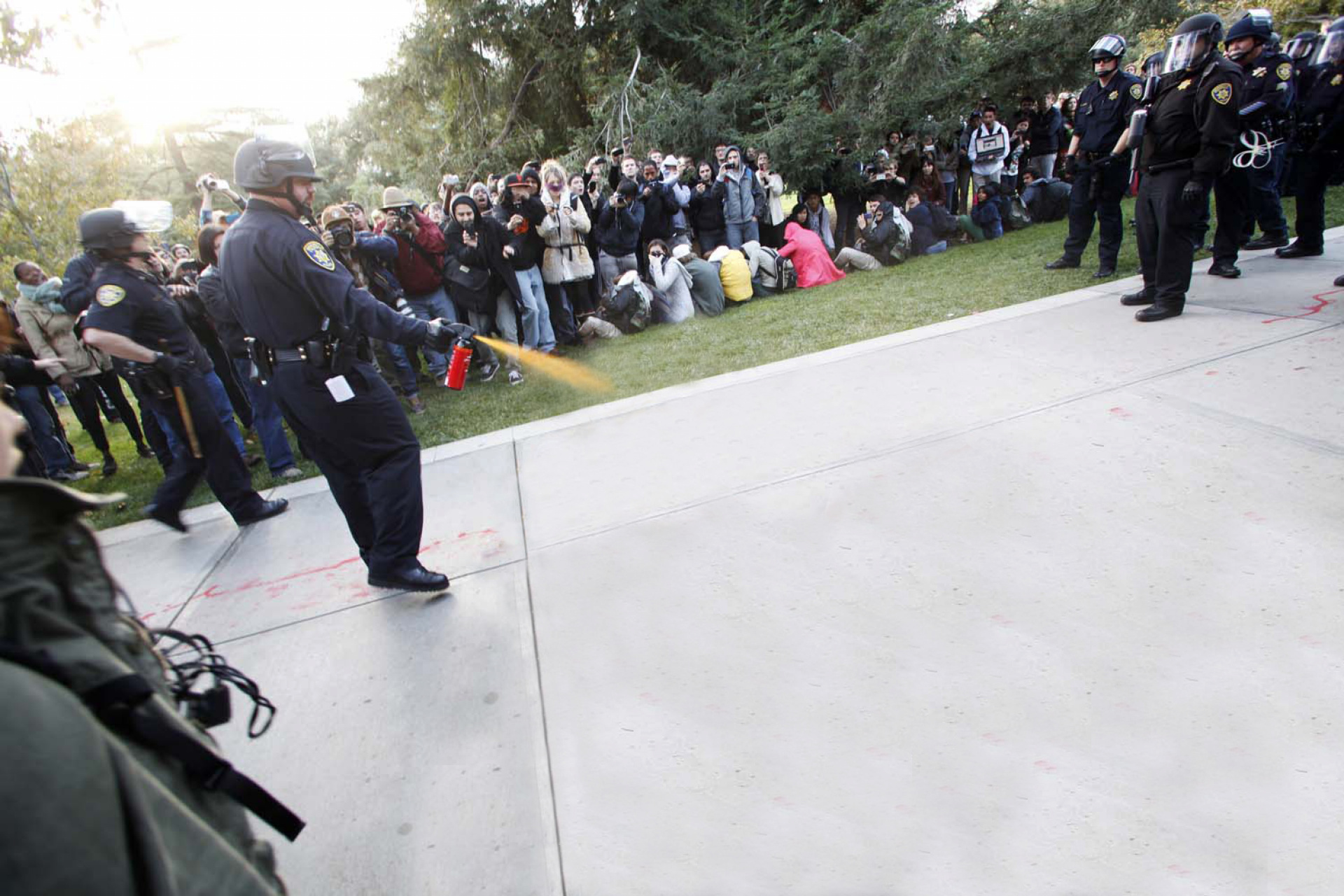 Police officer sprays pepper spray downwards, in front of onlookers.