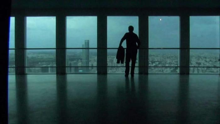 A man stands by large windows, gazing out over a sprawling cityscape.